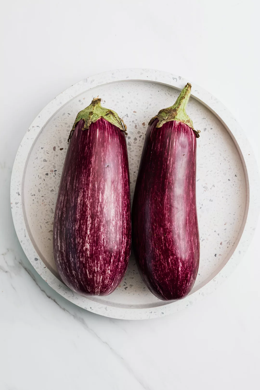 two fresh eggplants on a white plate on a marble background
